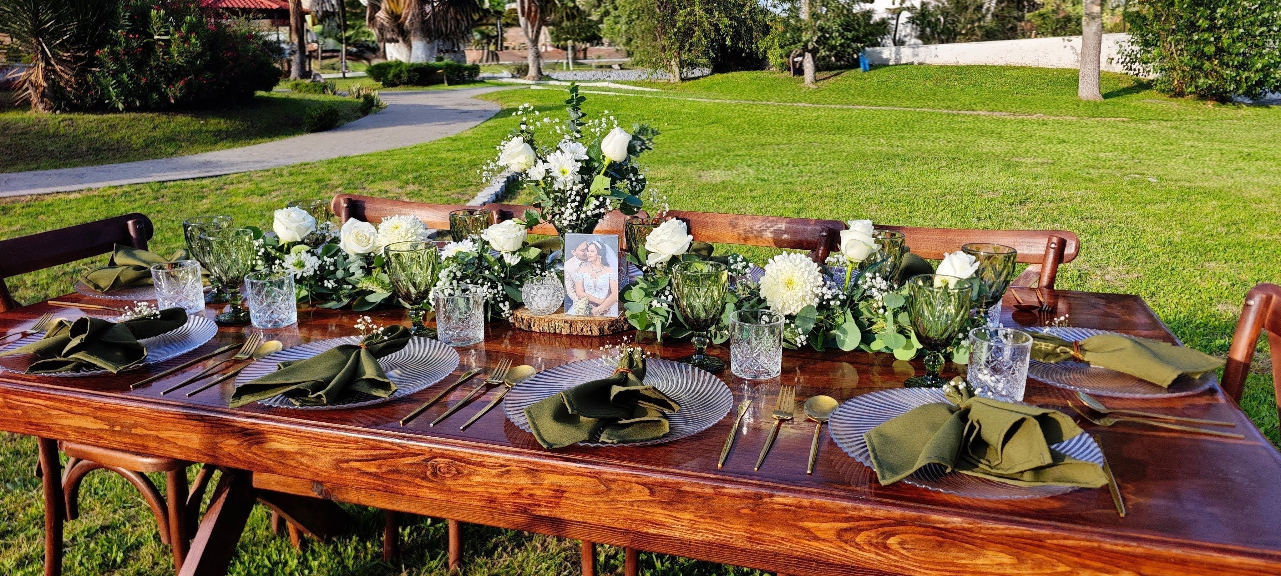 Mesa larga de madera al aire libre con flores blancas, cristalería verde y sillas crossback