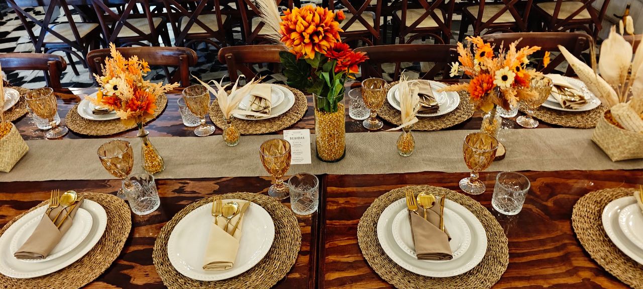 Mesa de madera con flores naranjas, cristalería ámbar, textiles beige y sillas crossback