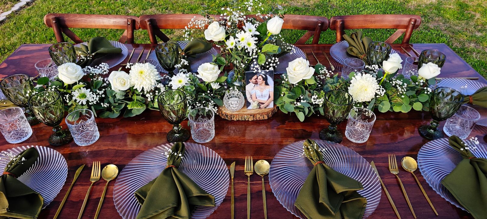 Mesa larga de madera con sillas crossback, flores blancas y cristalería verde en un jardín